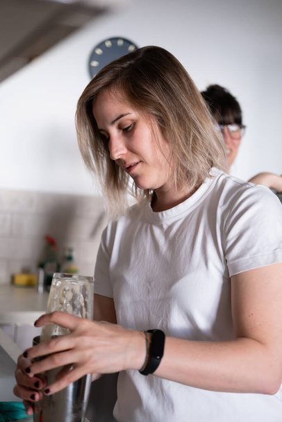A lesbian couple preparing homemade cocktails with a cocktail shaker in the kitchen - Photo, Image