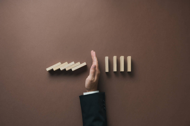 Top view of a hand of a businessman stopping falling dominos in a conceptual image of crisis prevention and solution. Over brown background. - Photo, Image