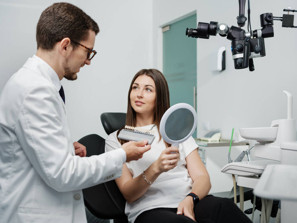 Молодой стоматолог в белом халате показывает палитру цвета эмали для красивой пациентки, сидящей в стоматологическом отделении. Устное эстетическое лечение. - Фото, изображение