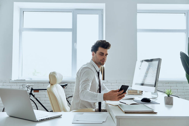 Confident man in shirt and tie looking at his smart phone while sitting in office - Photo, Image