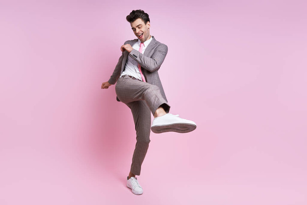 Happy young man in full suit throwing leg kick while standing against pink background - Photo, image