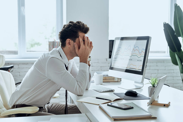 Depressed young man covering face with hands while sitting at his working place in office - Foto, Imagem