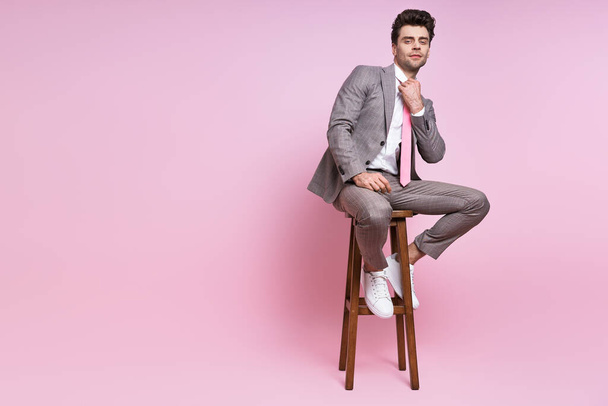 Confident man in full suit adjusting his necktie while sitting on the chair against pink background - Fotoğraf, Görsel