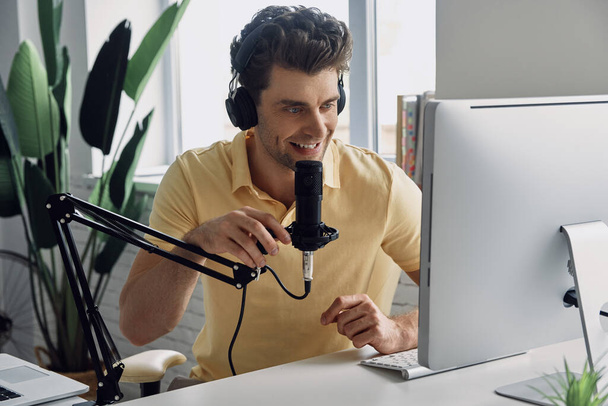 Confident young man using microphone and smiling while recording podcast in studio - Foto, Bild