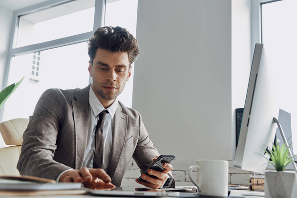 Confident young man using digital tablet and holding smart phone while sitting in office - Фото, изображение
