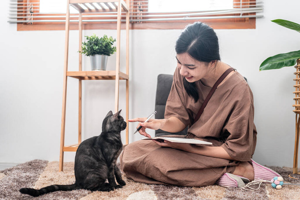 Красива жінка пише нотатки на блокноті і грає з прекрасною кішкою, сидячи, щоб розслабитися на килимі у вітальні
. - Фото, зображення