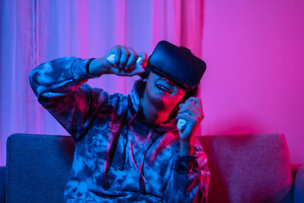 Young man wearing virtual reality (VR) glasses and uses joystick to play games with fun playing games in a room with red and blue lights, 3D game, Future games, Gadgets, Technology, Game concept.  - Foto, afbeelding