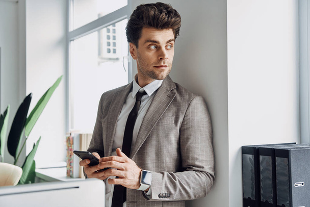 Handsome young man holding smart phone and looking through a window in office - Foto, Bild