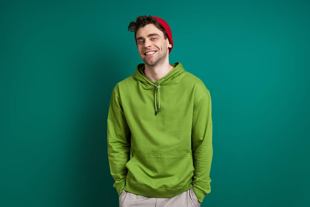Confident young man looking at camera and smiling while standing against green background - 写真・画像
