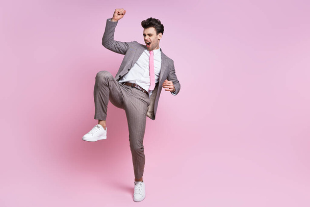 Excited young man in full suit gesturing while standing against pink background - Photo, image