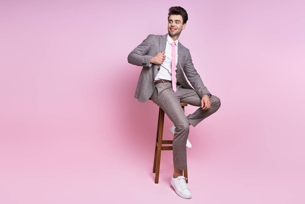 Handsome young man in full suit adjusting his jacket while sitting on chair against pink background - Фото, зображення