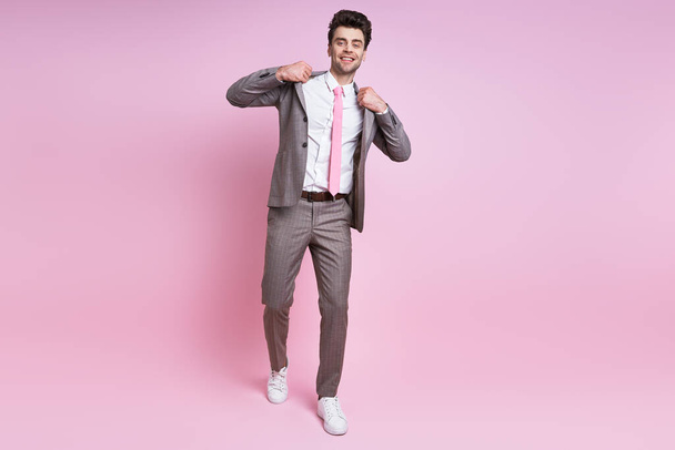 Cheerful young man in full suit adjusting his jacket while standing against pink background - Фото, зображення