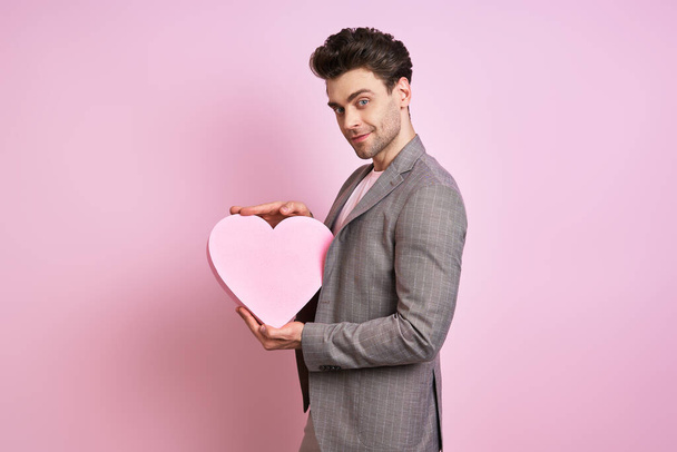 Happy young man in suit holding paper heart while standing against pink background - Фото, изображение