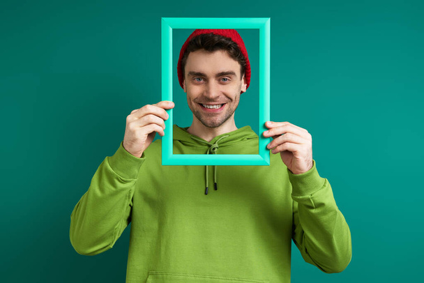 Handsome man looking through a picture frame and smiling while standing against green background - Valokuva, kuva