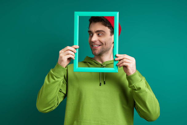 Playful young man looking through a picture frame while standing against green background - Photo, image