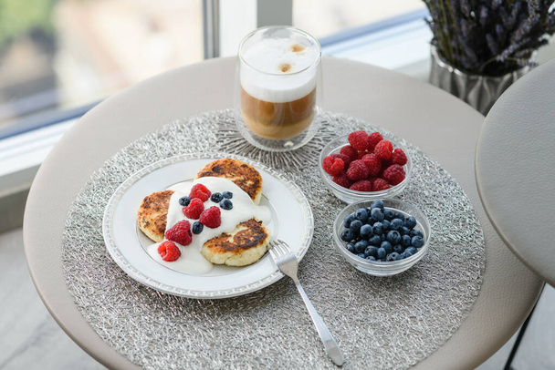 Cottage cheese pancakes for breakfast with blueberries and raspberries and sour cream. Cup of latte with delicious breakfast. Table with breakfast by the window on the background of the city. - Foto, immagini
