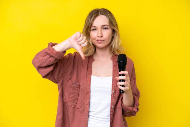 Молода співачка біла жінка підбирає мікрофон ізольовано на жовтому тлі, показуючи великий палець вниз з негативним виразом
 - Фото, зображення