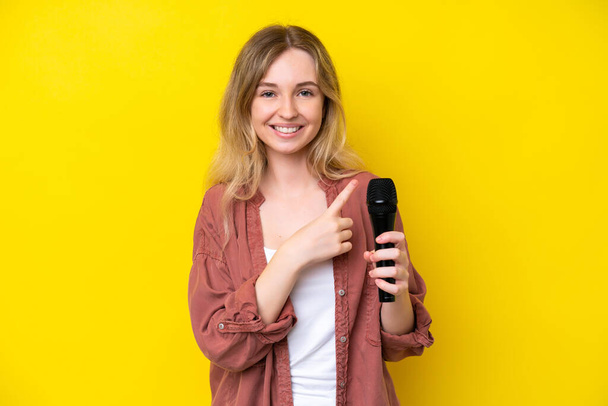 Young singer caucasian woman picking up a microphone isolated on yellow background pointing to the side to present a product - 写真・画像