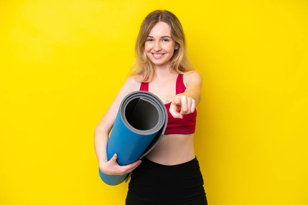 Young sport caucasian woman going to yoga classes while holding a mat isolated on yellow background pointing front with happy expression - 写真・画像