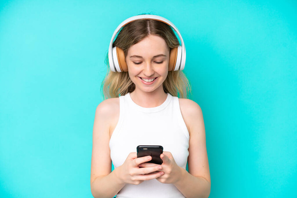 Blonde English young girl isolated on blue background listening music and looking to mobile - Foto, immagini
