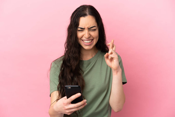 Young caucasian woman isolated on pink background using mobile phone with fingers crossing - Foto, afbeelding