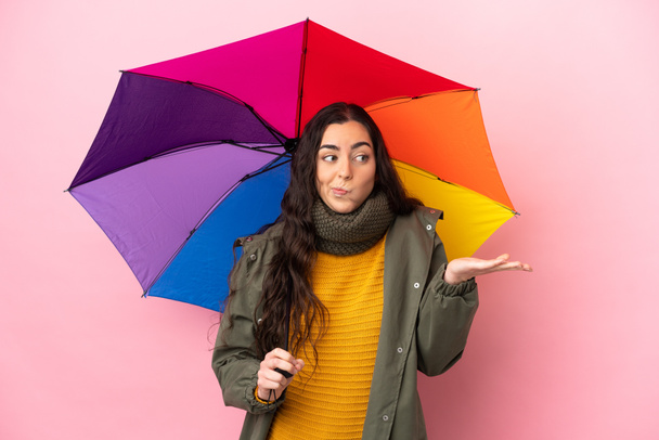 Young woman holding an umbrella isolated on pink background having doubts while raising hands - 写真・画像