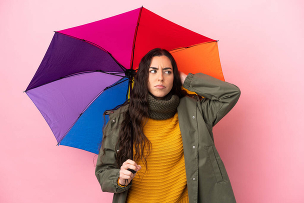 Mladá žena drží deštník izolovaný na růžovém pozadí s pochybnostmi - Fotografie, Obrázek