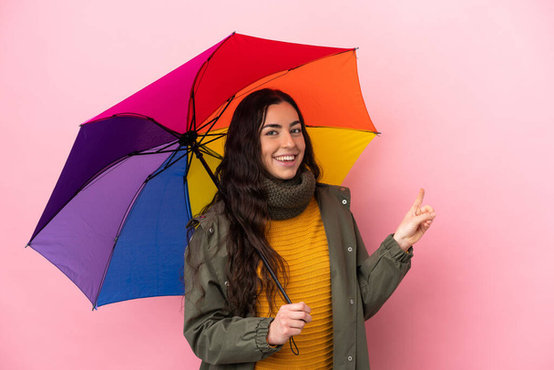 Young woman holding an umbrella isolated on pink background pointing back - Foto, Bild