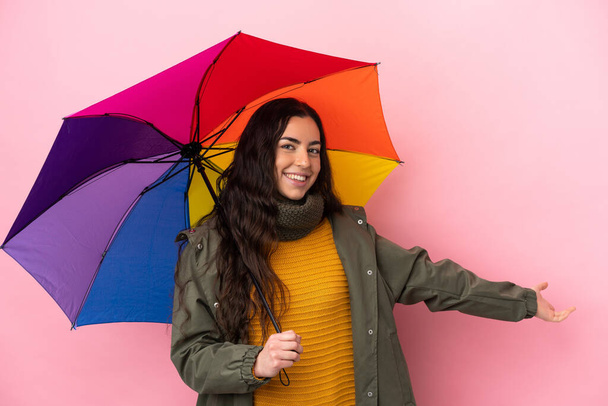 Young woman holding an umbrella isolated on pink background extending hands to the side for inviting to come - Fotoğraf, Görsel