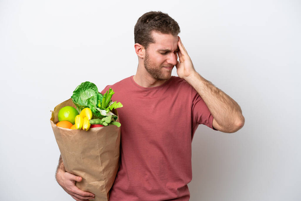 Young caucasian man holding a grocery shopping bag isolated on white background with headache - Foto, imagen