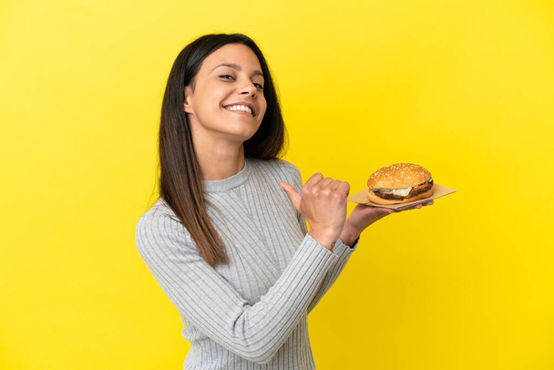 Young caucasian woman holding a burger isolated on yellow background proud and self-satisfied - Foto, Bild