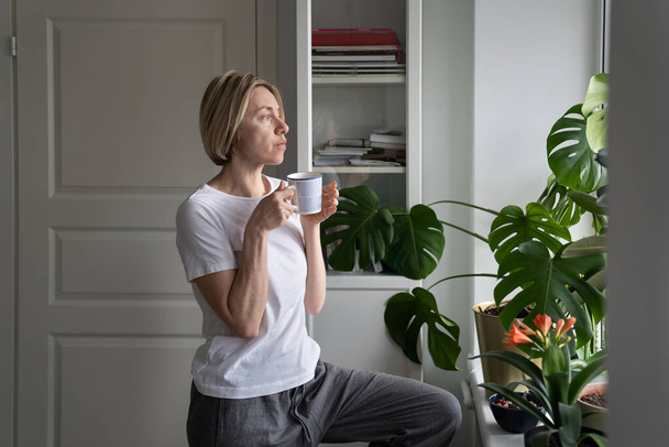 Tranquil middle-aged female enjoys hot drink from mug standing at windowsill. Mature blonde woman enjoys view outside window drinking hot coffee. Lady thinks about lifestyle and forthcoming day - Foto, Imagem