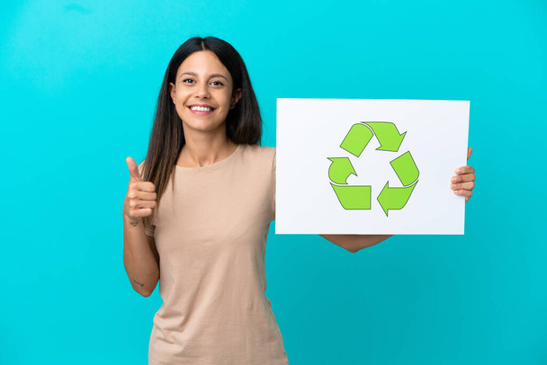 Young woman over isolated background holding a placard with recycle icon with thumb up - Photo, image
