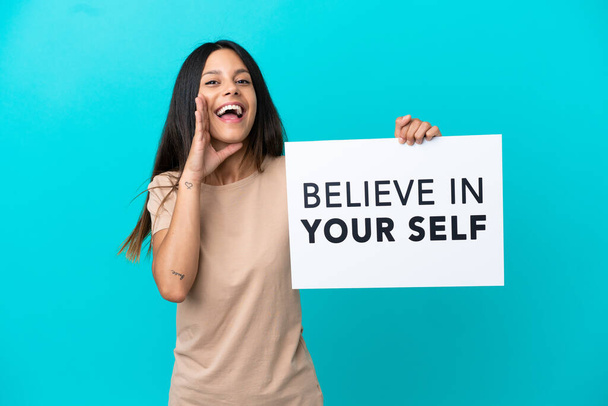 Νεαρή γυναίκα πάνω από απομονωμένη φόντο κρατώντας μια πινακίδα με κείμενο Πιστέψτε στον εαυτό σας και φωνάζοντας - Φωτογραφία, εικόνα