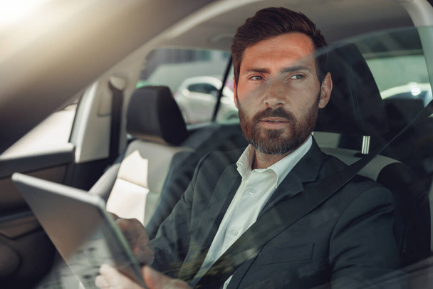 Бізнесмен працює на цифровому планшеті, сидячи на водійському сидінні в машині. Розмитий фон
 - Фото, зображення