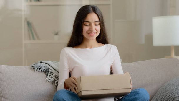 Surprised arab latin woman satisfied customer shopaholic sits on couch at home open delivery parcel postal carton box unpack present gift look at shipping goods purchases from internet clothing store - Photo, image