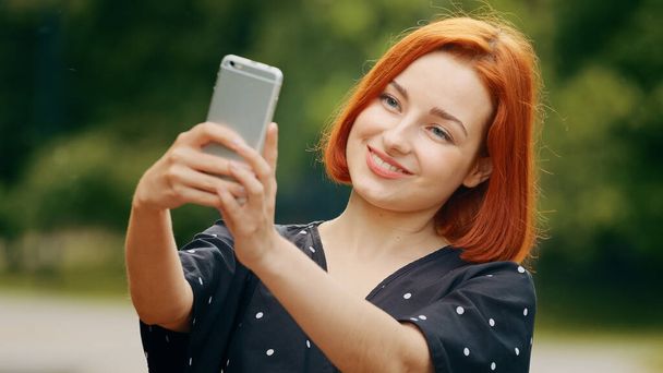 Beautiful caucasian redhead girl stylish model woman female blogger takes selfie photo self-admiration records video lifestyle blog on modern smartphone camera provides online live translation in park - Foto, immagini