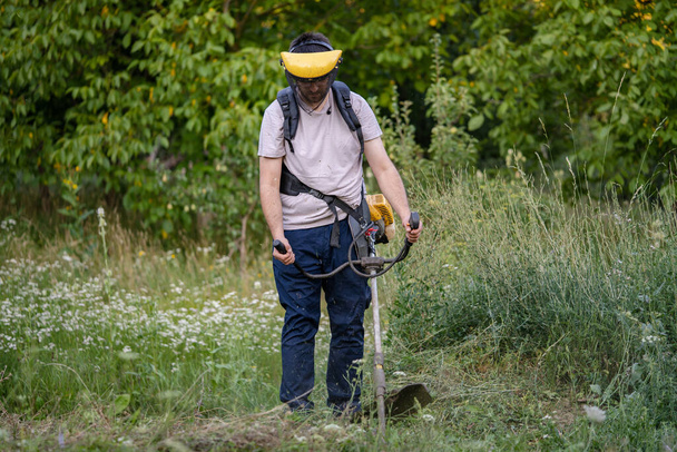 Молодой кавказский фермер-садовник, стоящий в поле с бензиновым триммером, подстригал траву, работая на поле, в солнечный день на деревенском переднем плане. - Фото, изображение