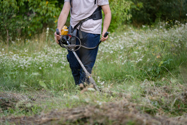 Молодой кавказский фермер-садовник, стоящий в поле со стринговым бензином, подстригал траву, работая на ферме в поле в солнечный день в середине деревни. - Фото, изображение