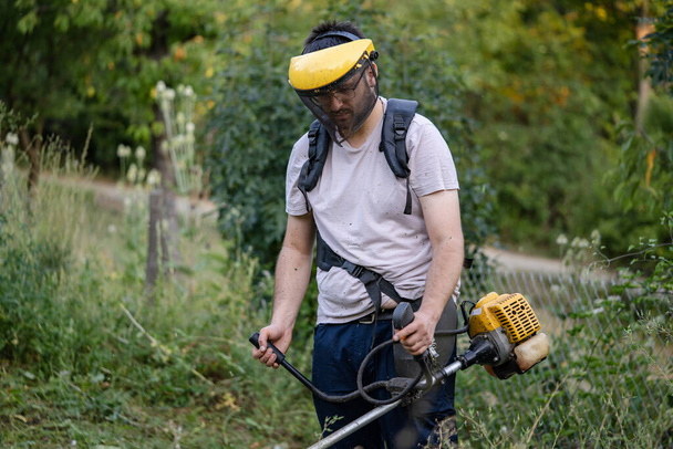 Молодий кавказький фермер садівник, що стоїть на полі, з бензиновим тримером.
 - Фото, зображення