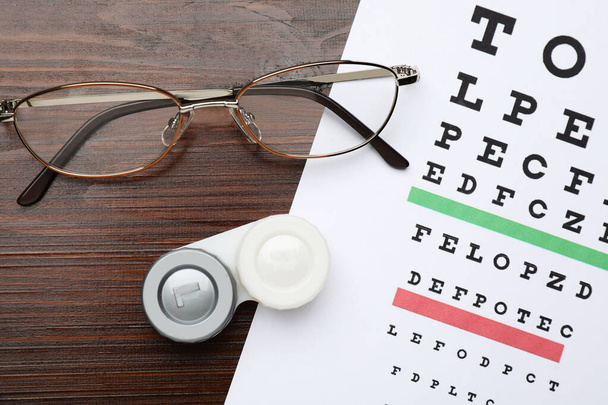 Case with contact lenses, glasses and eye chart test on wooden table, flat lay - Foto, afbeelding