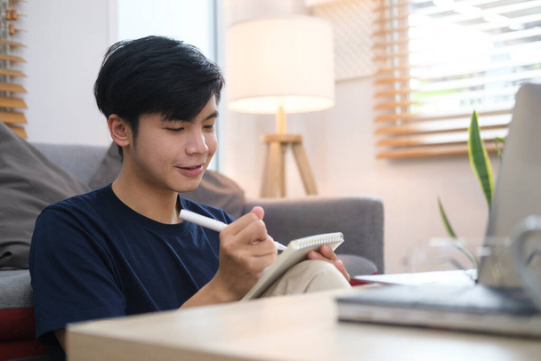 Young asian man sitting in bright living room and working online with laptop. - Photo, image