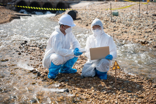 Ecologist sampling taken dead fish to inspaction and save data to laptop computer, Biologist wear protective suit and mask collects sample of waste water from industry, problem environment - Foto, Imagen