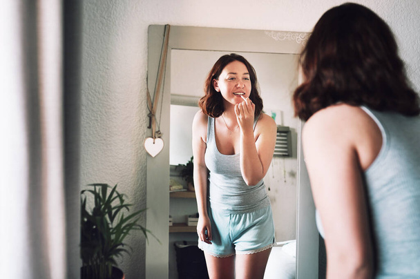 Looks like this colour will look good on me. an attractive young woman applying lipstick while standing in front of the mirror in her bedroom at home - Foto, imagen