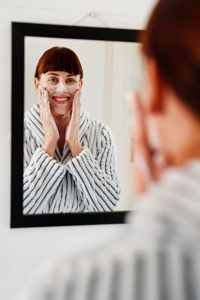 Shes sticking to this routine because its just perfect. an attractive young woman applying beauty treatment on her face while standing in front of the mirror at home - Fotoğraf, Görsel