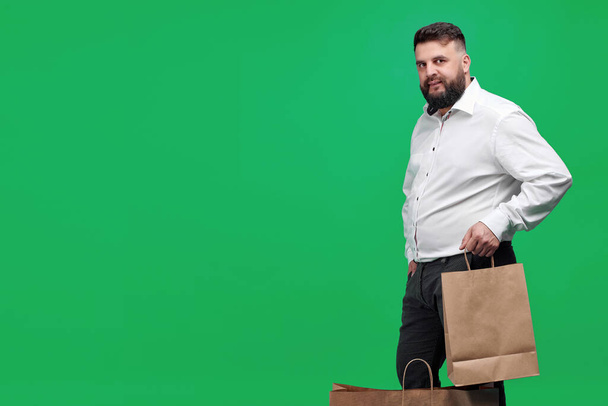 A Caucasian-looking man with a brown paper bag on a green background. A man with a white shirt with shopping bags. Studio shot of a man with purchases. - Foto, Imagen