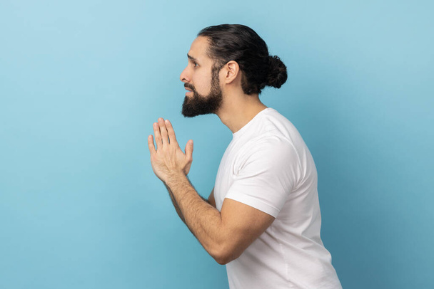 Please, I'm begging forgive. Side view of upset worried man with beard wearing white T-shirt looking with imploring desperate grimace, praying for help. Indoor studio shot isolated on blue background. - Fotografie, Obrázek