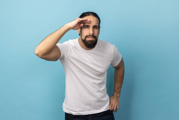 Portrait of curious man with beard wearing T-shirt keeping palm over head and looking attentively far away, peering with expectation at long distance. Indoor studio shot isolated on blue background. - Foto, Bild