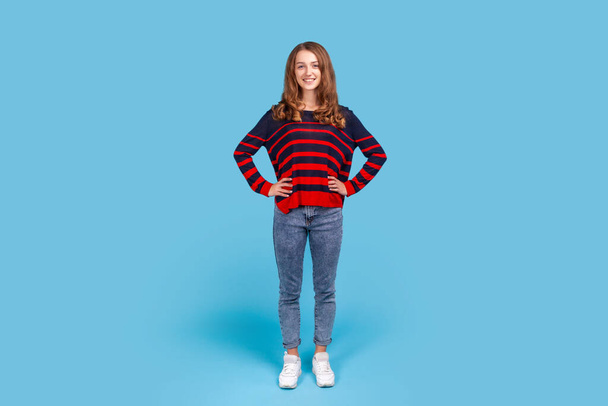 Full length portrait of positive happy woman wearing striped casual style sweater and jeans standing with hands on hips, looking at camera. Indoor studio shot isolated on blue background. - Photo, image