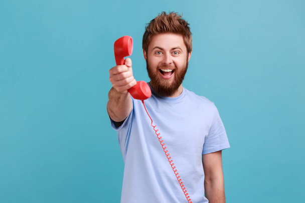 Portrait of smiling positive bearded man holding in hands red retro telephone, waiting for your call, looking at camera with excited expression. Indoor studio shot isolated on blue background. - Foto, Imagem